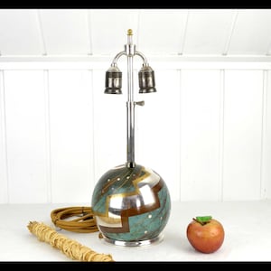 May include: A vintage table lamp with a spherical base featuring a teal, gold, and silver geometric design. The lamp has a silver metal stem and two bulb sockets. An apple and a bundle of wrapped sticks are next to the lamp.
