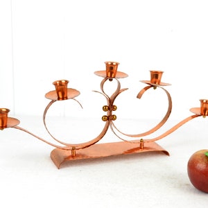 May include: A copper candle holder with five candle cups. The holder is shaped like a series of curved lines and has a hammered finish.