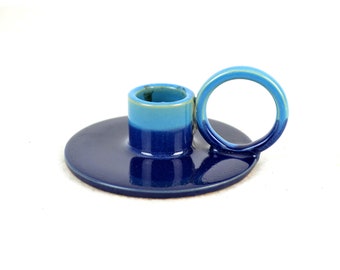 May include: A blue and teal ceramic candle holder with a ring handle. The holder has a round base and a cylindrical top.