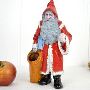 Könnte beinhalten: Eine Vintage-Figur von Weihnachtsmann mit einem rot-weißen Anzug, einem grauen Bart und einem Sack in der Hand. Die Figur ist aus bemaltem Gips und hat einen rustikalen Look.
