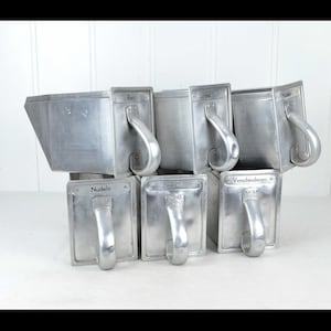 Peut inclure: Ensemble d'ustensiles de cuisine vintage en aluminium, comprenant des récipients rectangulaires avec des poignées incurvées et des étiquettes de texte. Les outils sont empilés, mettant en valeur leur design industriel. Les étiquettes indiquent "Nudeln" et "Zwiebeln".
