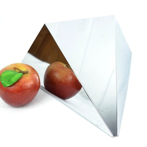 May include: A silver, triangular prism sits on a white surface. Two red apples with green leaves are in front of the prism. The prism reflects the apples.