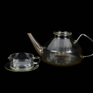 May include: A clear glass teapot and a matching cup and saucer. The teapot has a round body and a curved spout. The cup has a handle and a saucer with a raised edge.