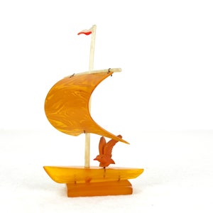 May include: A small, orange, wooden sailboat with a white sail and a red and white flag. A small, orange, wooden bird is perched on the deck.