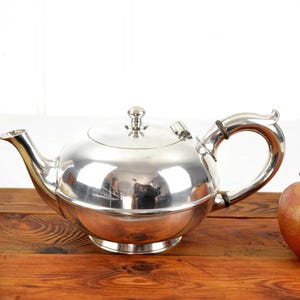 Op de afbeelding: Een glanzende zilveren theepot met een gebogen tuit en handvat, en een deksel met een kleine knop. De theepot staat op een houten oppervlak naast een rode appel. Het is een keukenartikel.
