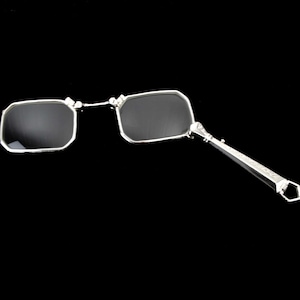 Könnte beinhalten: Silbergerahmte, rechteckige Brille mit dunklen Gläsern. Die Brille hat einen dekorativen Griff mit geometrischem Design. Der Hintergrund ist einfarbig schwarz.