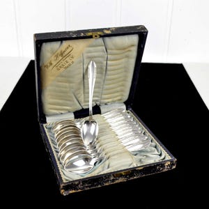 Könnte beinhalten: Ein antikes Silberlöffel-Set in einer schwarzen Box mit hellblauem Satinfutter. Das Set enthält einen großen Servierlöffel und mehrere kleinere Löffel, alle mit aufwendigen Details. Auf der Box steht "Wilh. Hoffmann, GREIZ I.V."