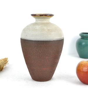 Puede incluir: Un jarrón de cerámica marrón y blanco con una superficie texturizada. El jarrón tiene una boca ancha y un cuello estrecho.