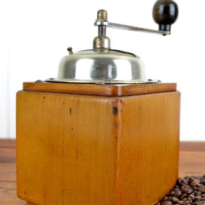 K&ouml;nnte beinhalten: Eine antike Kaffeem&uuml;hle aus Holz mit silbernem Oberteil und Kurbel. Die M&uuml;hle steht auf einer Holzoberfl&auml;che mit Kaffeebohnen, die um sie herum verstreut sind.