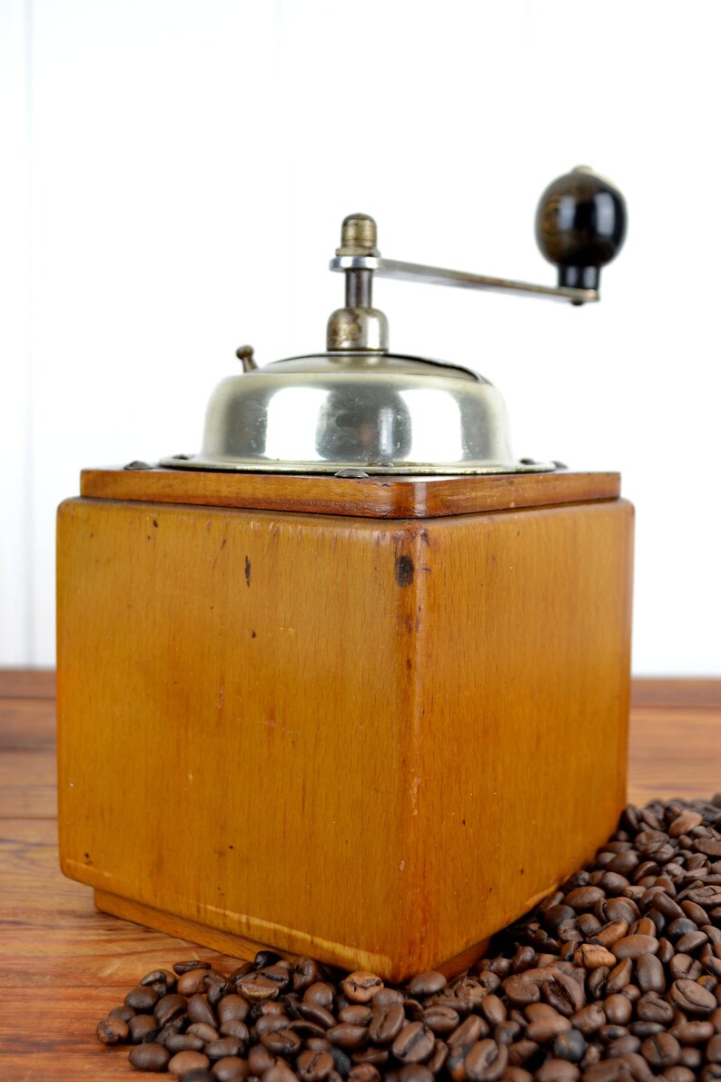 K&ouml;nnte beinhalten: Eine antike Kaffeem&uuml;hle aus Holz mit silbernem Oberteil und Kurbel. Die M&uuml;hle steht auf einer Holzoberfl&auml;che mit Kaffeebohnen, die um sie herum verstreut sind.