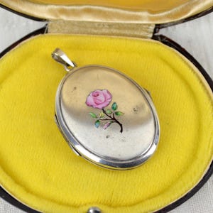 May include: Silver oval locket with a pink rose design, resting on a yellow velvet interior within a dark brown case. The locket is suspended from a silver bail. The case is open, revealing the locket's details.