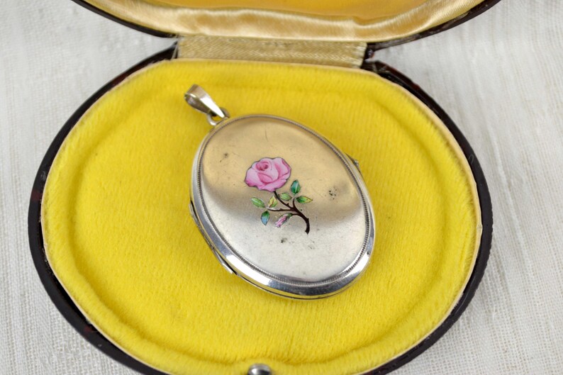 May include: Silver oval locket with a pink rose design, resting on a yellow velvet interior within a dark brown case. The locket is suspended from a silver bail. The case is open, revealing the locket's details.