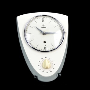 May include: A white wall clock with a shield-shaped frame and a circular face featuring the Junghans logo, black numerals, and silver hands. Below the clock face is a cream-colored rotary timer dial with black markings ranging from 0 to 60.