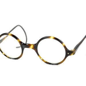 Puede incluir: Gafas redondas con un patrón de carey. Las monturas son una mezcla de marrón y negro, con lentes transparentes. Las patillas se curvan alrededor de las orejas. Las gafas están sobre un fondo blanco.