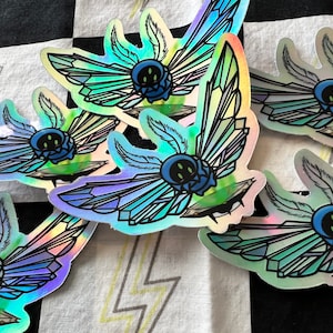 May include: Five iridescent holographic stickers featuring a blue creature with three eyes and crystal wings. The creature is sitting on a green platform.