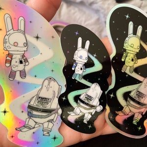 Può includere: Tre adesivi olografici con un coniglio astronauta in una scena spaziale. Il coniglio astronauta indossa una tuta spaziale bianca con una visiera nera e ha orecchie lunghe. La scena spaziale presenta una galassia vorticosa con stelle e una cometa.