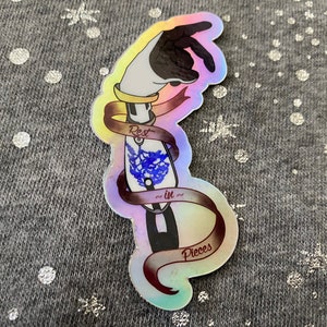 May include: A holographic sticker with a rainbow iridescent background. The sticker features a black hand holding a blue and white object with a ribbon wrapped around it. The ribbon has the text "Rest in Pieces" on it.