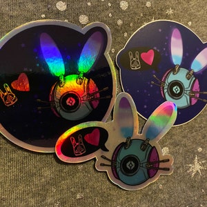 May include: Three holographic stickers featuring a cartoon robot with bunny ears and a heart. The robot is blue, white, and black. The stickers are on a gray fabric background.