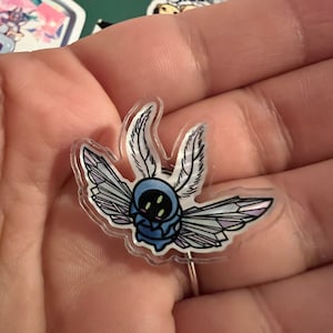 May include: A clear acrylic pin featuring a stylized blue creature with feathery antennae and geometric wings. The creature has four green eyes and a blue body. The wings have a gradient of purple and white.