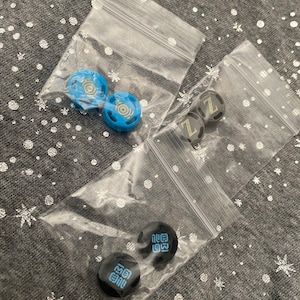 May include: Three small plastic bags containing different colored and patterned round buttons. The buttons are black with white lettering, blue with white lettering, and black with blue lettering.