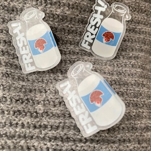 Può includere: Tre spille in plastica trasparente a forma di bottiglie di latte con un'etichetta blu e bianca e la scritta "FRESH!" stampata su di esse. L'etichetta presenta un'immagine di un fungo rosso e marrone.