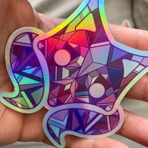 May include: A holographic sticker with a geometric design in shades of purple, blue, and red. The sticker has a star-shaped outline and a unique, abstract pattern.