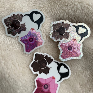 May include: Three holographic stickers featuring a geometric design with a central circular element. Each sticker has a speech bubble with a heart. The stickers are in shades of brown, pink, and silver.