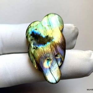 Op de afbeelding: Een gepolijste labradoriet edelsteen gesneden in een abstracte vorm. De steen vertoont iriserende kleuren van blauw, groen, geel en paars. De edelsteen wordt vastgehouden in een witte gehandschoende hand.