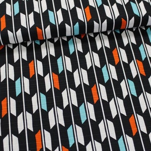 Könnte beinhalten: Schwarzer Stoff mit einem sich wiederholenden Muster aus weißen, orangen und türkisfarbenen geometrischen Formen. Der Stoff ist in der Mitte gefaltet und ein Holzlineal ist im Hintergrund sichtbar.