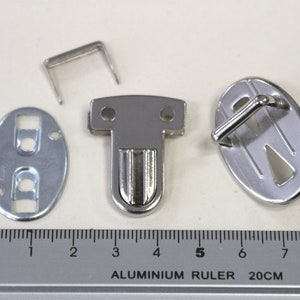 Könnte beinhalten: Ein Satz silberfarbener Metallverschlüsse für eine Tasche oder Handtasche. Das Set enthält einen Haken, einen Riegel, eine Platte mit zwei Löchern und eine kleine Metallschlaufe. Die Verschlüsse sind neben einem in Zentimetern markierten Lineal abgebildet.