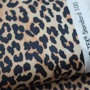 Könnte beinhalten: Nahaufnahme einer Stoffrolle mit Leopardenmuster. Der Stoff hat einen beigefarbenen Hintergrund mit dunkelbraunen Flecken. Der Rand der Stoffrolle zeigt den Text "OEKO-TEX® Standard 100".