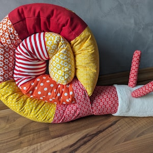 Könnte beinhalten: Ein farbenfrohes, Patchwork-Kissen in Schneckenform mit einer rot-weiß-gelb gestreiften Schale. Das Kissen ist aus weichem Stoff gefertigt und perfekt zum Kuscheln.