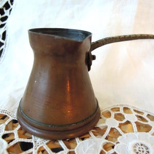 May include: A copper-colored Turkish coffee pot with a long, textured handle. The pot has a wide base that tapers to a narrow neck and a flared rim. The handle is attached to the side of the pot. The pot is sitting on a white surface.