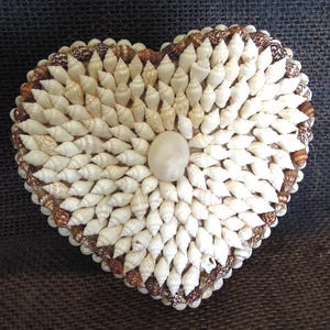 May include: Heart-shaped decorative box covered in small, white seashells. The box is edged with brown and white shells, and a large, smooth, off-white stone is in the center. The box is on a dark, textured surface.