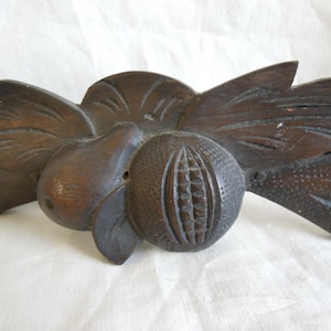 May include: A dark brown, carved wooden decorative piece featuring a central sphere with vertical grooves, flanked by a pear-shaped element and stylized leaves. The piece has a rustic, antique appearance.