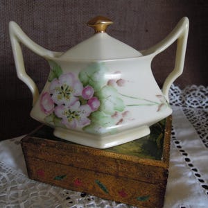 May include: A white porcelain sugar bowl with a gold lid and floral design. The bowl is sitting on a wooden box with a gold finish.