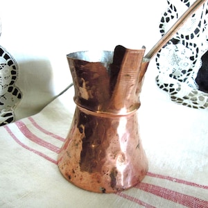 May include: A hammered copper coffee pot with a wide base, a narrow neck, and a long metal handle. The exterior surface features a textured, reflective finish with visible indentations from the hammering process, and the interior is lined in silver.