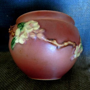 May include: A small, round, antique-style ceramic pot in a muted rose color. The pot is adorned with a raised floral design featuring white and yellow flowers with green leaves, and brown stems. The pot has a slightly textured surface.