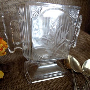 May include: Clear glass sugar bowl with etched fern design and two handles. The bowl has a rectangular base and is accompanied by several silver spoons. The background is a textured brown surface.