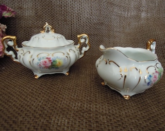 May include: A white porcelain sugar bowl and creamer set with gold trim and floral accents. The sugar bowl has a lid and both pieces have delicate floral designs in pink, yellow, and blue.