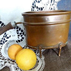 May include: A copper-colored, vintage cooking pot with a wooden handle and three small legs. Two oranges and a lemon sit in front of the pot on a white lace doily. The pot has a round shape and a slightly flared rim.