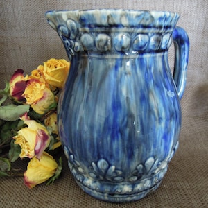 May include: A blue and white ceramic pitcher with a decorative design. The pitcher has a wide mouth and a handle. The pitcher is sitting on a burlap surface.
