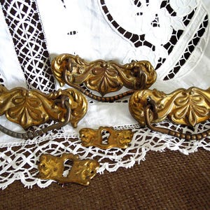 May include: Four antique brass drawer pulls with ornate floral designs. The pulls have a chain-like detail and are arranged on a white lace background.