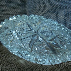 May include: A clear, oval-shaped glass bowl featuring an intricate, multi-faceted geometric pattern. The surface is covered in a combination of starburst motifs, cross-hatching, and diamond-shaped cuts that reflect light across the entire piece.
