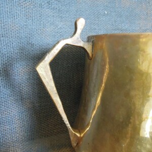 May include: Close-up of a brass mug with a unique handle design. The handle is shaped like a figure with outstretched arms. The mug has a hammered texture and a warm, golden color, suggesting it is vintage or handmade.