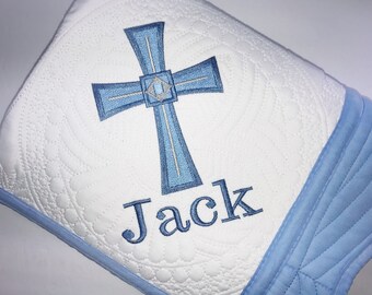 Könnte beinhalten: Eine weiße gesteppte Babydecke mit einem blauen Kreuz und dem Namen "Jack", der darauf gestickt ist. Die Decke hat einen blauen Rand.