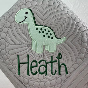 May include: A gray baby blanket with a green dinosaur applique and the name "Heath" embroidered in green.