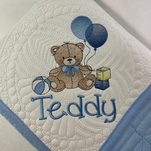 May include: A white quilted baby blanket with a blue border. The blanket features an embroidered brown teddy bear holding a blue and white ball, two blue balloons, and a yellow block. The text "Teddy" is embroidered below the bear.