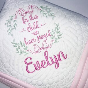 May include: White baby quilt with pink trim and embroidered text. The quilt features a floral wreath design with the words "for this child we have prayed" and the name "Evelyn" in pink script.