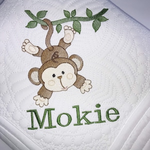 Könnte beinhalten: Eine weiße gesteppte Babydecke mit einem braunen Affen, der an einer grünen Rebe hängt. Der Affe hat einen braunen Schwanz und lächelt. Die Decke ist mit dem Namen "Mokie" in grüner Stickerei personalisiert.